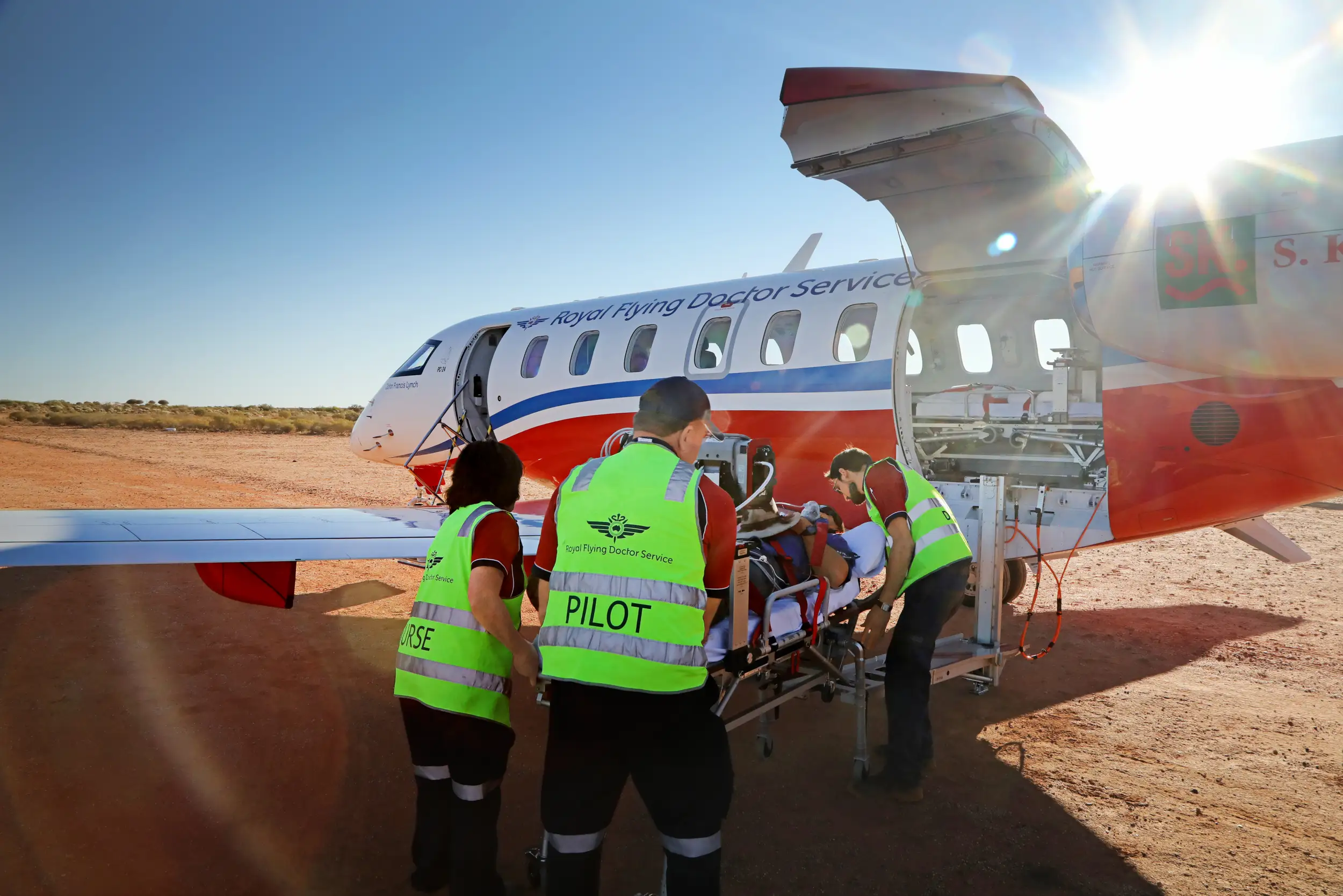 A PC-12 RFDS aircraft in the wilderness, as paramedics carefully load a patient on a stretcher into the plane.