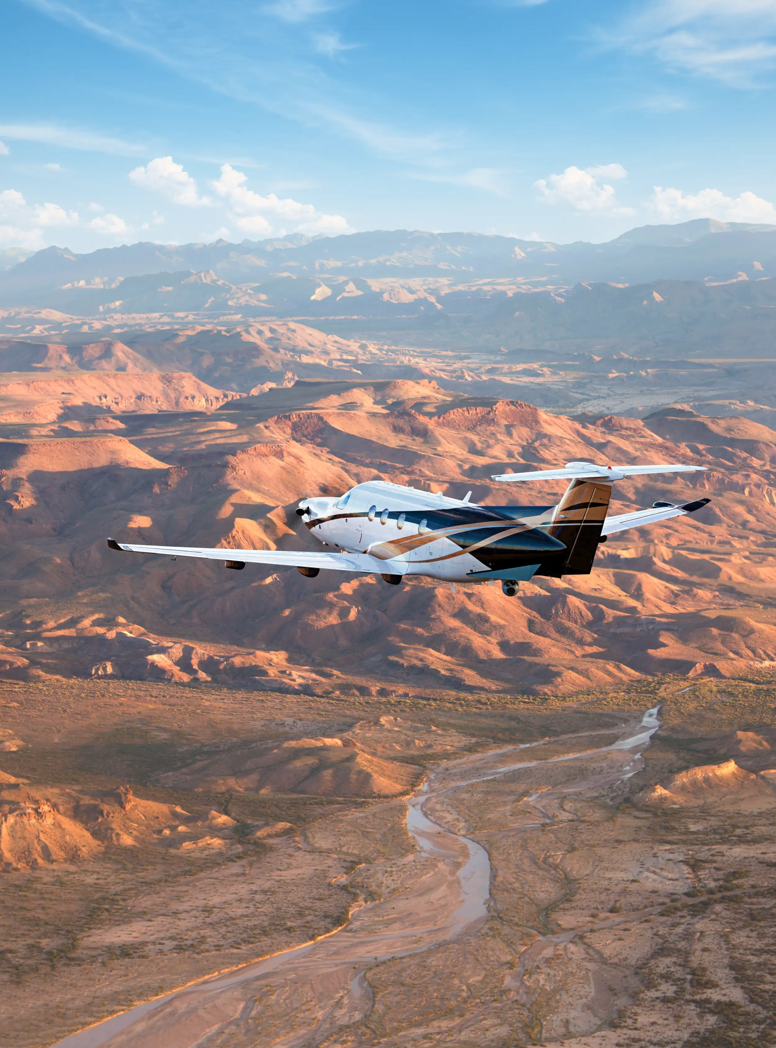 A PC-12 Spectre flies over reddish mountains with a blue sky.